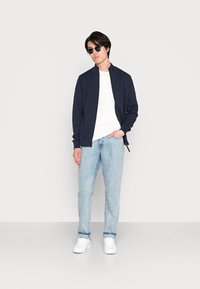 Marineblauwe zip-up jas over een wit T-shirt, gecombineerd met lichtblauwe jeans en witte sneakers. Model draagt ronde zonnebril.