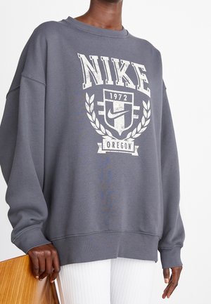 Osoba nosící volnou tmavě šedou mikinu Nike s bílým logem "1972 Oregon" a vavřínovým věncem, držící dřevěnou židli.