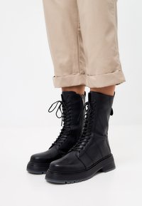 Botas de combate de cuero negro con suela gruesa, cordones en la parte delantera y detalle de cremallera. Textura suave con acentos de costura reforzada.