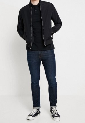 Veste bombardier noire avec fermeture éclair blanche, polo noir, jean bleu foncé et baskets noires avec accents blancs. Look décontracté et monochrome.