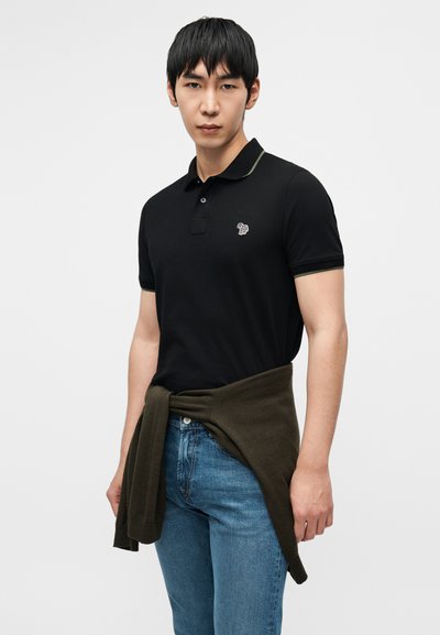 Μαύρο πόλο μπλουζάκι με κοντά μανίκια, αντιθετικό γιακά, κεντημένο λογότυπο, συνδυασμένο με μπλε τζιν και ελιά πράσινο πουλόβερ δεμένο γύρω από τη μέση.