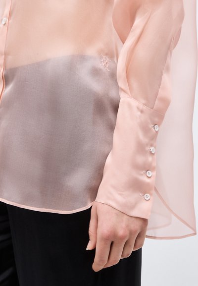 Απαλή πορτοκαλί μπλούζα με μακριά μανίκια, μανσέτες με κουμπιά και κεντημένο λογότυπο στο στήθος, φορεμένη με μαύρο παντελόνι.