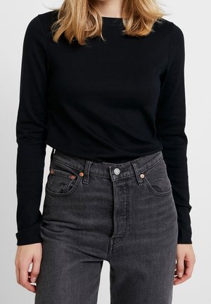 Personne portant une chemise noire à manches longues rentrée dans un jean denim gris foncé taille haute, debout devant un fond uni.