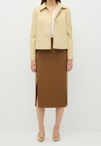 Giacca beige a forma accorciata con colletto, abbinata a una gonna a tubino marrone con spacchi laterali. Scarpe in tonalità chiara con dettagli dorati.