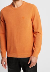 Pull en tricot orange avec une finition texturée, doté d'un col rond et de poignets et ourlets côtelés. Inclut un petit logo brodé sur la poitrine.