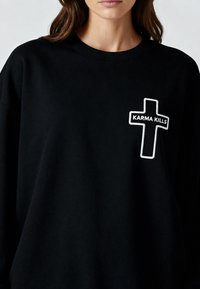 Femme portant un sweat-shirt noir avec une croix blanche en contour et le texte "KARMA KILLS" sur la poitrine.