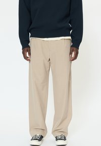 Pantaloni beige a gamba larga con tessuto strutturato, caratterizzati da pieghe laterali. Abbinati a un maglione di lana blu navy e sneakers nere.
