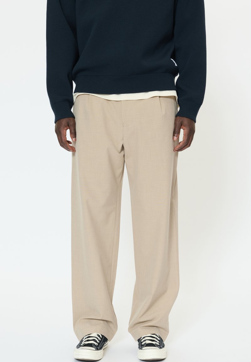 Pantaloni beige a gamba larga con tessuto strutturato, caratterizzati da pieghe laterali. Abbinati a un maglione di lana blu navy e sneakers nere.