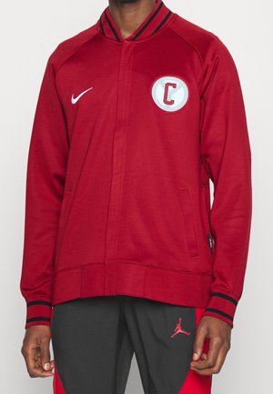 Chaqueta roja con cremallera y cuello acanalado, que presenta un parche circular blanco con una "C." Fabricada con un tejido suave y con puños a rayas azul marino.