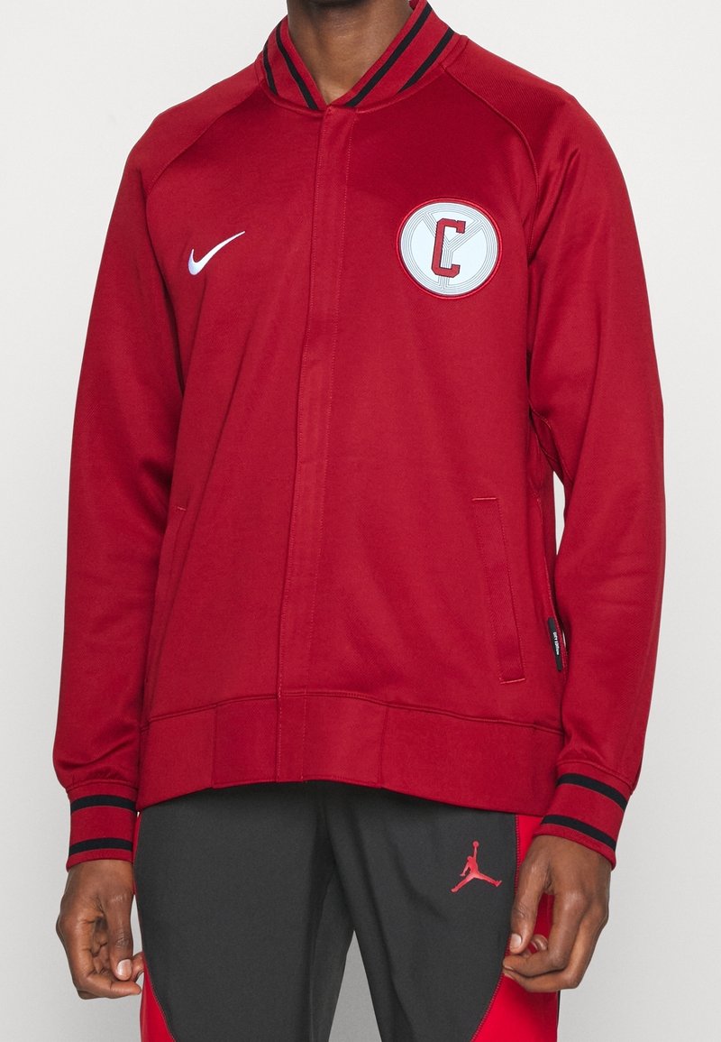 Rød zip-jakke med ribbet krave, der har et hvidt cirkulært patch med et "C." Fremstillet af glat stof med søm i marineblå.