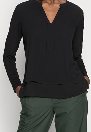 Personne portant un haut noir à manches longues avec un petit col en V et un pantalon vert foncé, les mains placées de manière décontractée dans les poches.