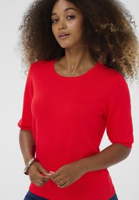 Rød strikket kortærmet top med rund halsudskæring. Glat tekstur og en tætsiddende silhuet, parret med et sølvarmbånd på håndleddet.