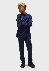 Marinblå träningsdräkt med zip-up tröja och matchande byxor, med en logotyp på bröstet och sidochevron-accenter, tillverkad av ett slätt, atletiskt material.