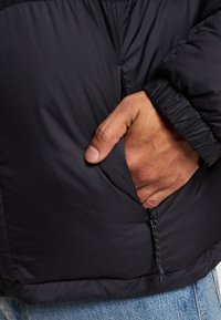 Veste noire isolée avec une poche latérale zippée. Le tissu est lisse, avec un léger brillant et des poignets élastiques.
