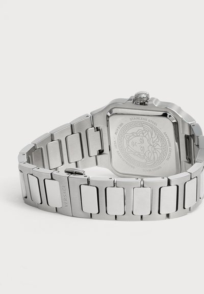 Montre-bracelet en acier inoxydable argenté avec un bracelet à maillons et un logo Méduse gravé au dos du boîtier carré de la montre.