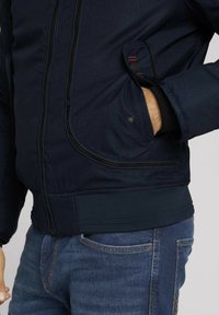 Giacca imbottita blu navy con chiusura a zip, tessuto testurizzato, tasche laterali, polsini elastici e orlo a costine.