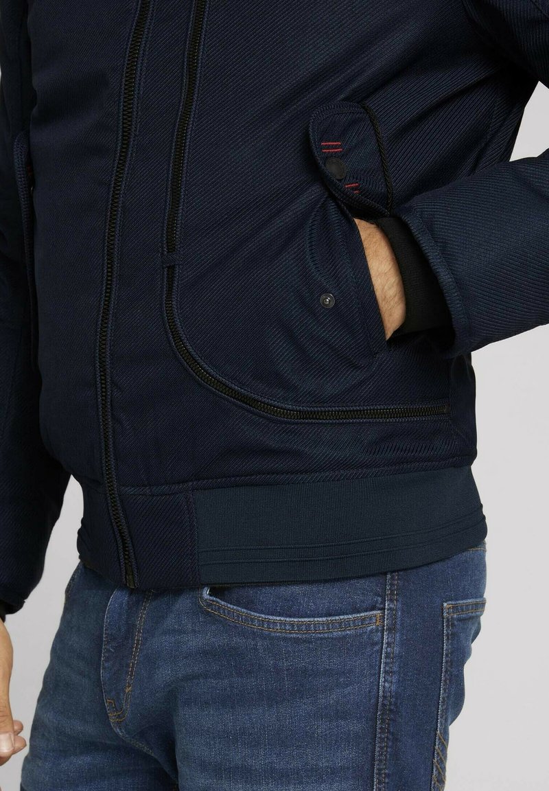 Giacca imbottita blu navy con chiusura a zip, tessuto testurizzato, tasche laterali, polsini elastici e orlo a costine.