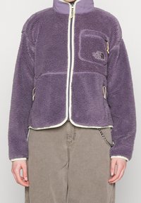 Giacca in pile viola con zip frontale, colletto alto e rifiniture bianche. Presenta una tasca sul petto con zip e dettagli con cordoncino.