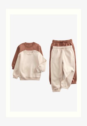 Lille barn står iført en beige sweatshirt og joggingbukser med sorte og hvide sneakers på en ensfarvet lys baggrund.