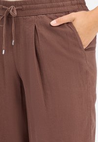 Bruine, lichte broek met een elastische tailleband, voorplooien en zijzakken. De stof heeft een gladde textuur en een subtiele glans.