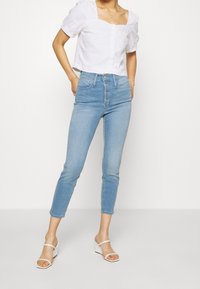 Jeans azul claro de cintura alta con una silueta ajustada, cierre de cinco botones y largo recortado. Combinados con una blusa blanca de manga corta.