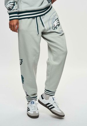 Helle graue Sweatpants mit grünen und weißen Streifen, ausgestattet mit Logos und Designs von Sportteams. Kombiniert mit schwarz-weißen Sneakers.
