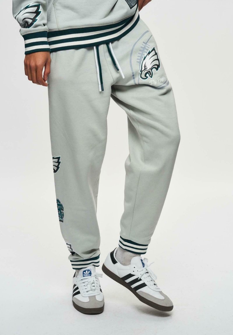 Lysgrå træningsbukser med grønne og hvide striber, prydet med sportsholdets logoer og designs. Sammen med sorte og hvide sneakers.