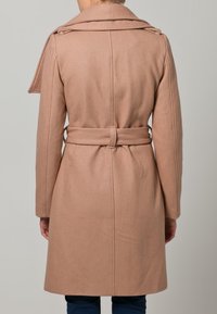 Ljusbeige ullkappa med bälte i midjan, långa ärmar och breda slag; har en slät textur och rak skärning.