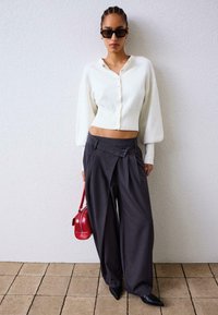 Biely skrátený kardigán s gombíkmi, šedé široké nohavice s bočným gombíkom a červená kabelka. Čierne špicaté topánky.