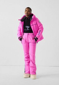 Persona che indossa pantaloni da sci rosa acceso e giacca imbottita, maglione nero con cerniera e grafica sciistica, in piedi contro uno sfondo uniforme.