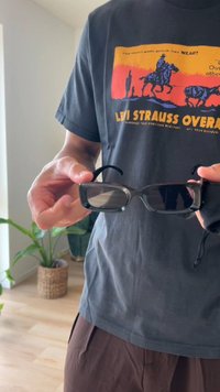 Óculos de sol pretos com armação retangular, acabamento brilhante e lentes escuras, segurados numa mão, com um estojo suave preso.