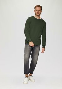 Dunkelgrüner Strickpullover mit rundem Ausschnitt, kombiniert mit grauen Jeans mit umgeschlagenen Säumen und weißen Sneakers, die ein dezentes Logo-Detail aufweisen.