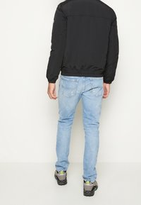 Giacca bomber nera imbottita, jeans slim blu chiaro e sneaker scure con suole macchiate, viste da dietro su uno sfondo semplice.