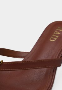 Καφέ δερμάτινο σανδάλι με λεπτό λουράκι και λεπτομέρεια χρυσής αγκράφας σε φωτεινό φόντο, εμφανίζοντας την επιφάνεια πατούσας και μέρος της σόλας.