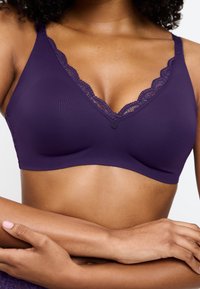 Violetti rintsikka, jossa on v-pukuinen muotoilu, sileä kangas, pitsireunus kaula-aukossa ja koristepainike keskellä.