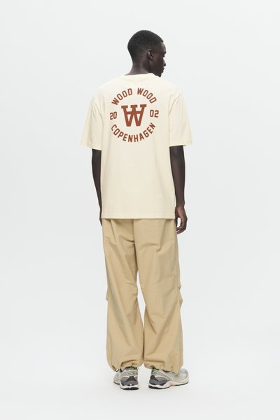Uomo in piedi con la schiena visibile, indossa pantaloni larghi beige, sneakers bianche e una t-shirt color crema con il testo e il logo "WOOD WOOD COPENHAGEN 2002".