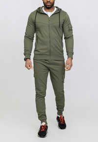 Survêtement de sport vert olive en tissu lisse, comprenant un sweat à capuche zippé, un pantalon cargo avec poches latérales et des baskets noires avec des accents orange.