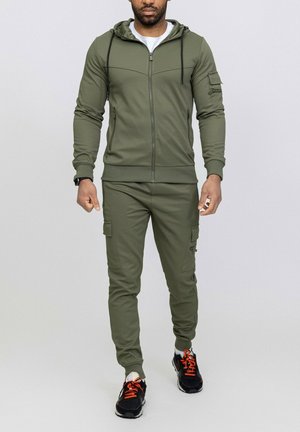Survêtement de sport vert olive en tissu lisse, comprenant un sweat à capuche zippé, un pantalon cargo avec poches latérales et des baskets noires avec des accents orange.