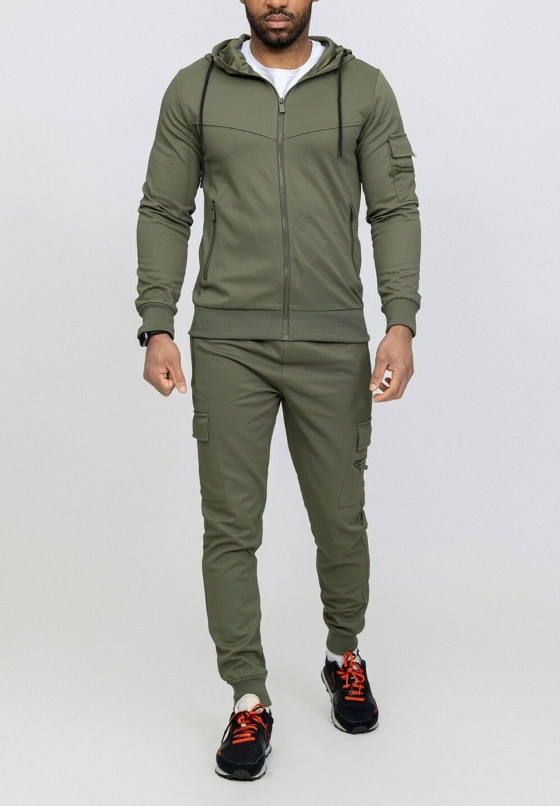 Survêtement de sport vert olive en tissu lisse, comprenant un sweat à capuche zippé, un pantalon cargo avec poches latérales et des baskets noires avec des accents orange.