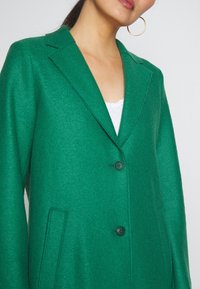 Blazer de mezcla de lana verde con un acabado ligeramente texturizado, solapas con muesca, dos botones negros y bolsillos laterales.