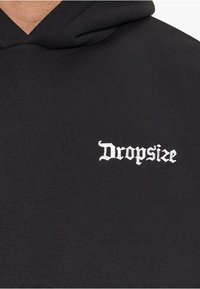 Schwarze Kapuzenjacke aus weichem Stoff mit weißen stickierten Text "Dropsize" in einer stilisierten Schriftart im Brustbereich.