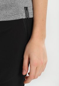 Tyylikkäät mustat urheilulegginsit yhdistettynä harmaaseen, raidalliseen lyhythihaiseen paitaan, jossa on teksturoitu etiketti sivussa. Lähikuva käden sijainnista.