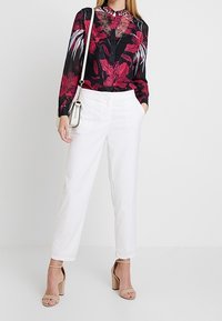 Schwarze Bluse mit rotem Blumenmuster, Bluse hat Knöpfe und einen Kragen; kombiniert mit weißen, taillierten Hosen und beigen, hochhackigen Sandalen.
