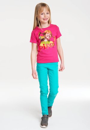 Helles pinkes T-Shirt mit einem bedruckten Design, das eine Figur und Text zeigt. Kombiniert mit türkisfarbenen Hosen und grauen Sneakers.