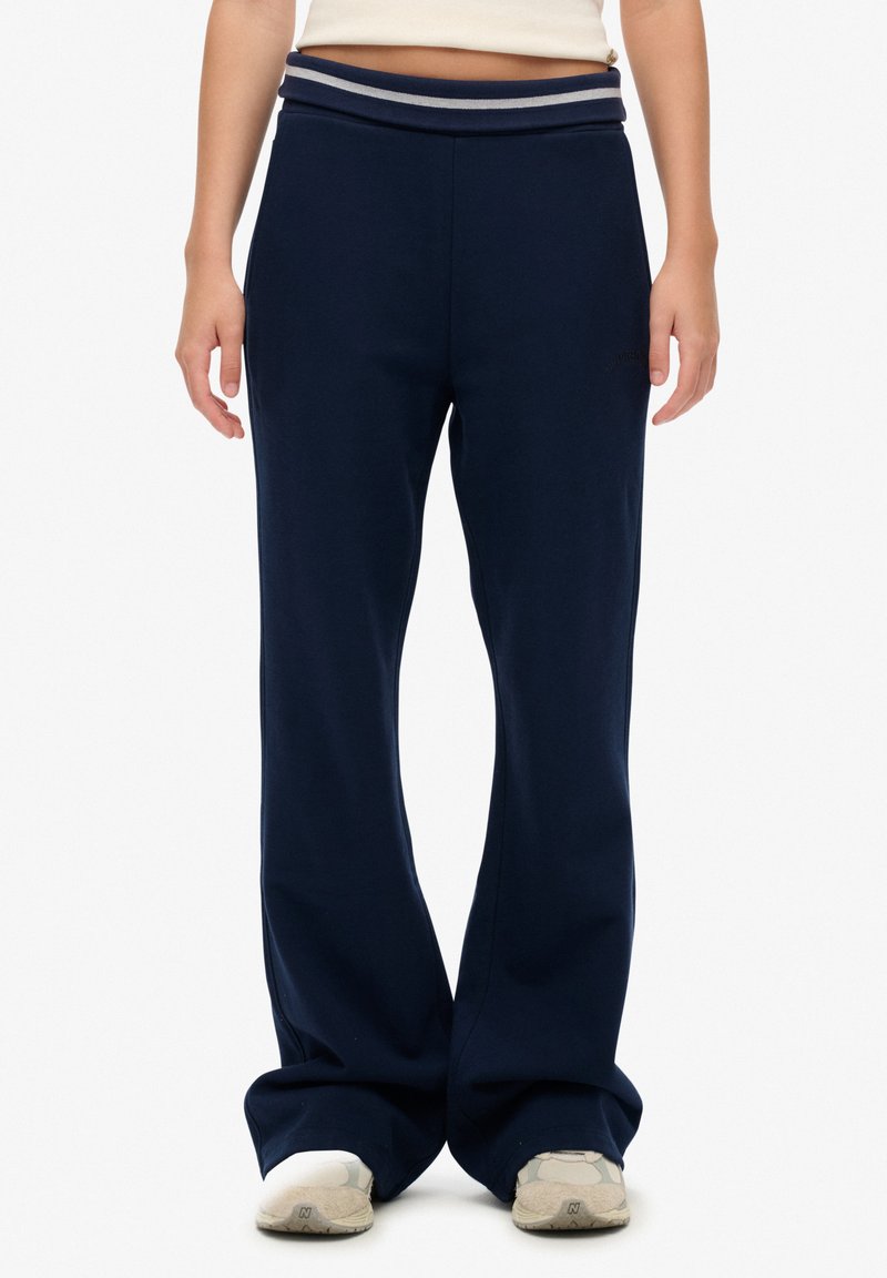 Personne portant un pantalon large bleu marine avec une ceinture rayée et des baskets blanches, debout devant un fond uni.