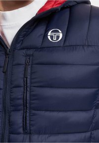 Marine pufferjakke med rød hette, med horisontal quilted design, glidelåslommer og en hvit logo på brystet.