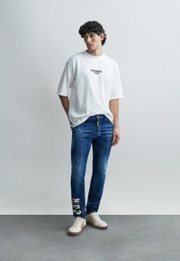 Weißes übergroßes T-Shirt mit Logo, blaue Jeans mit Used-Details und weiße Sneaker mit dezenten Akzenten. Einfaches, lässiges Outfit.