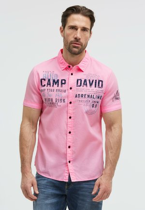 Man with short brown hair wearing a bright pink short-sleeve button-up shirt with black text and blue jeans, standing against a plain background.
