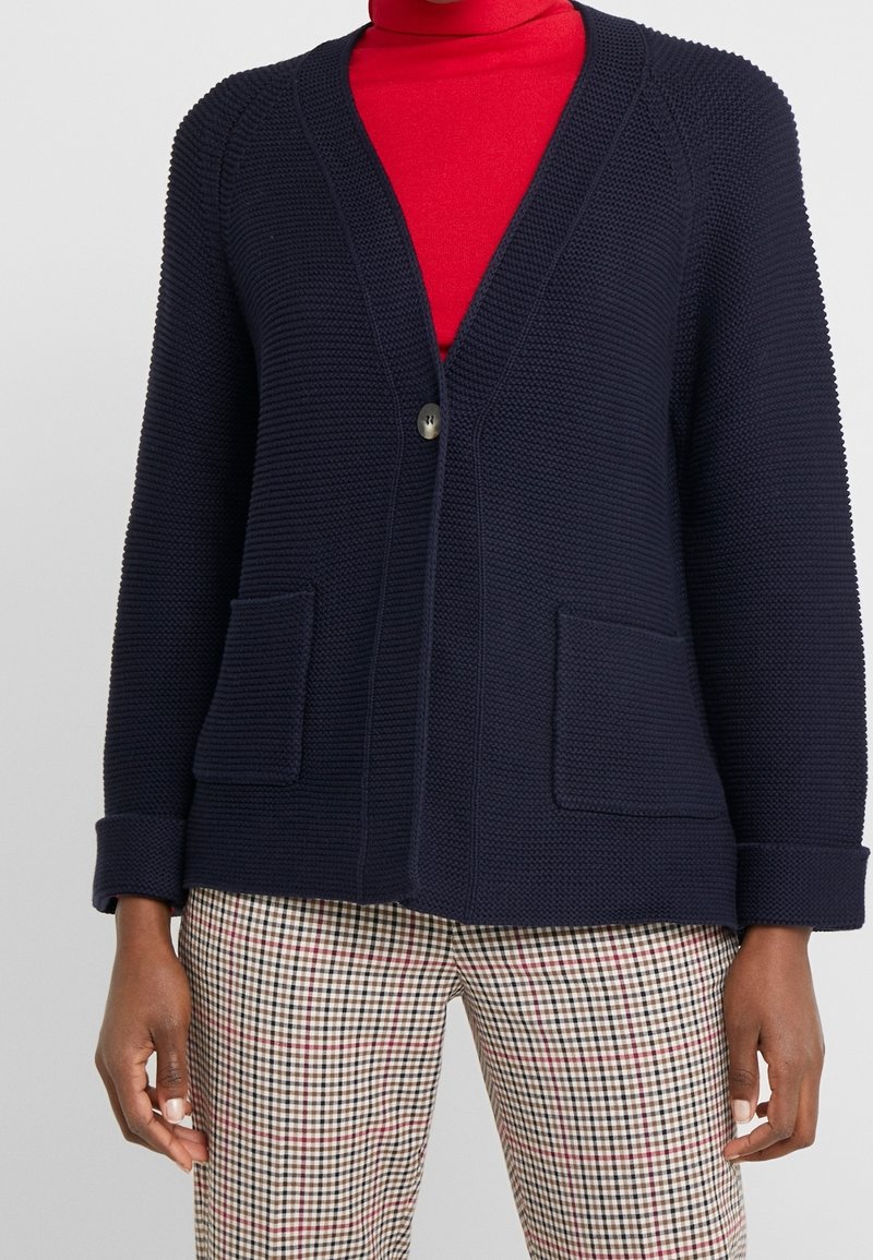 Cardigan lavorato a maglia blu navy con scollo a V, chiusura con un bottone singolo e due tasche frontali. Tessuto a trama con polsini arrotolati. Pantaloni a quadri sotto.