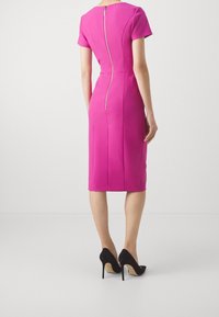 Femme debout portant une robe rose vif ajustée, mi-longue jusqu'aux genoux, avec une fermeture éclair apparente au dos, associée à des chaussures à talons hauts noires.